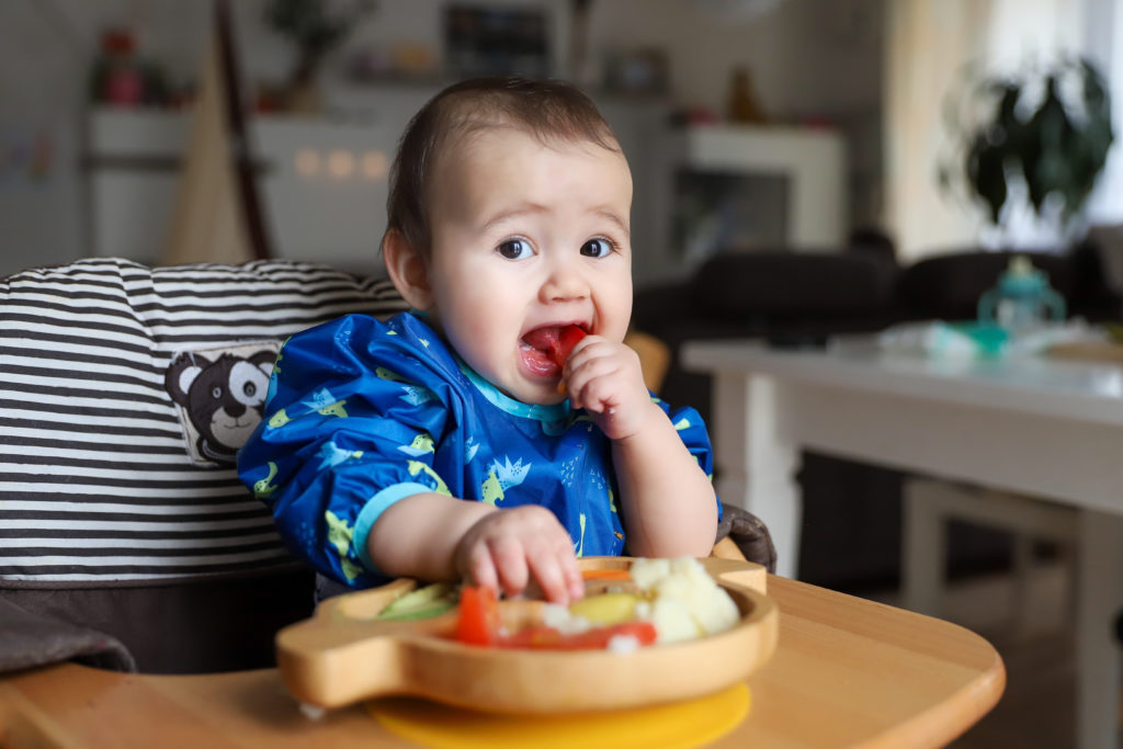 bebé subido en una trona comiendo comida verduras solo de un plato de madera